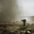 Valentino Bellini. Agbogbloshie, Accra, Ghana. A guy is standing in the midst of smoke, fire and residual parts of electronic equipment as he burns it to extract some copper contained inside to resell later to earn his daily food.